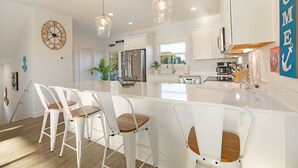 A modern kitchen with white cabinetry, a large island with bar stools, pendant lighting, and a decorative clock on the wall. - Decks with Ocean Views, Grill, 2 King Suites, Dogs Welcome, 2 Min Walk to Beach (Surf City)