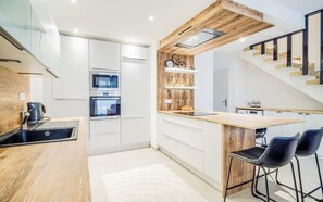 Une cuisine moderne avec des meubles blancs, un plan de travail en bois et une cuisinière intégrée. On y trouve une étagère en bois ornée d’objets décoratifs et un vélo.