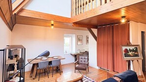 A spacious loft with a high ceiling, wooden beams, a dining area, a living room with a sofa and a painting, and a balcony with a railing. - Atelier d’Artiste – Ville Close à pied (Concarneau)