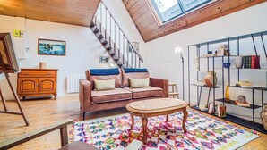 A loft-style room with a wooden ceiling, a staircase, a brown leather sofa, a colorful rug, and a bookshelf. - Atelier d’Artiste – Ville Close à pied (Concarneau)