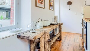 A rustic kitchen with wooden flooring, a wooden bench, and a window overlooking a brick building. - Atelier d’Artiste – Ville Close à pied (Concarneau)