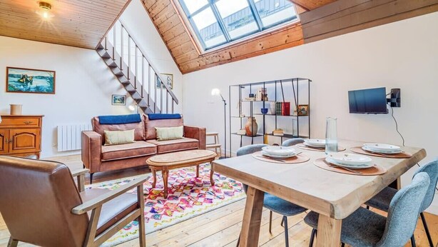 A spacious living area with a high ceiling, a wooden staircase, a sofa, a coffee table, a dining table set for four, and a bookshelf. - Atelier d’Artiste – Ville Close à pied (Concarneau)