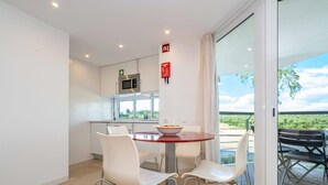 A modern kitchen with white cabinets and a microwave, a dining area with a red table and white chairs, and a large sliding glass door leading to an outdoor patio with a view of the countryside. - 🏖️ White Golden - AC - Wi-Fi - Access Pools & Beach Included (Cabanas de Tavira)