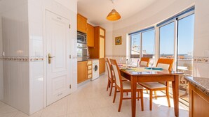 A modern kitchen with white cabinetry, a dining area with wooden chairs, and a large window offering a city view. - Tavira Getaway with Balcony and Sea View (Tavira)