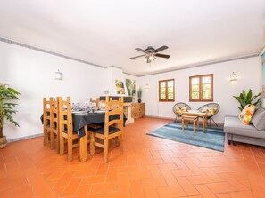 Un coin repas avec une table dressée pour quatre personnes, entourée de chaises en bois. Un ventilateur de plafond est installé au-dessus, et un canapé avec des coussins se trouve à l’arrière-plan.