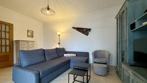 Ein Wohnzimmer mit grauem Sofa, einem kleinen Tisch, einer Lampe und einem Fernseher auf einem Schrank.