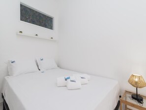 Un lit bien fait, avec du linge de lit blanc et des oreillers à rayures bleues, une petite table de chevet avec une lampe, et une étagère murale ornée d’objets décoratifs.