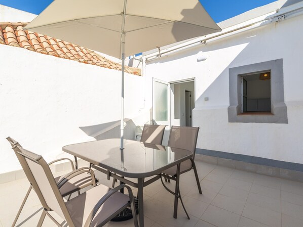 Une terrasse avec une table et des chaises en verre, un parasol beige et un toit recouvert de tuiles.