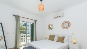 A bedroom with a bed, bedside tables, a bench, a window with curtains, and wall-mounted air conditioning. - Casa do Lagar - 4 mins walk to train station - quiet - wi-fi (Conceição de Tavira)