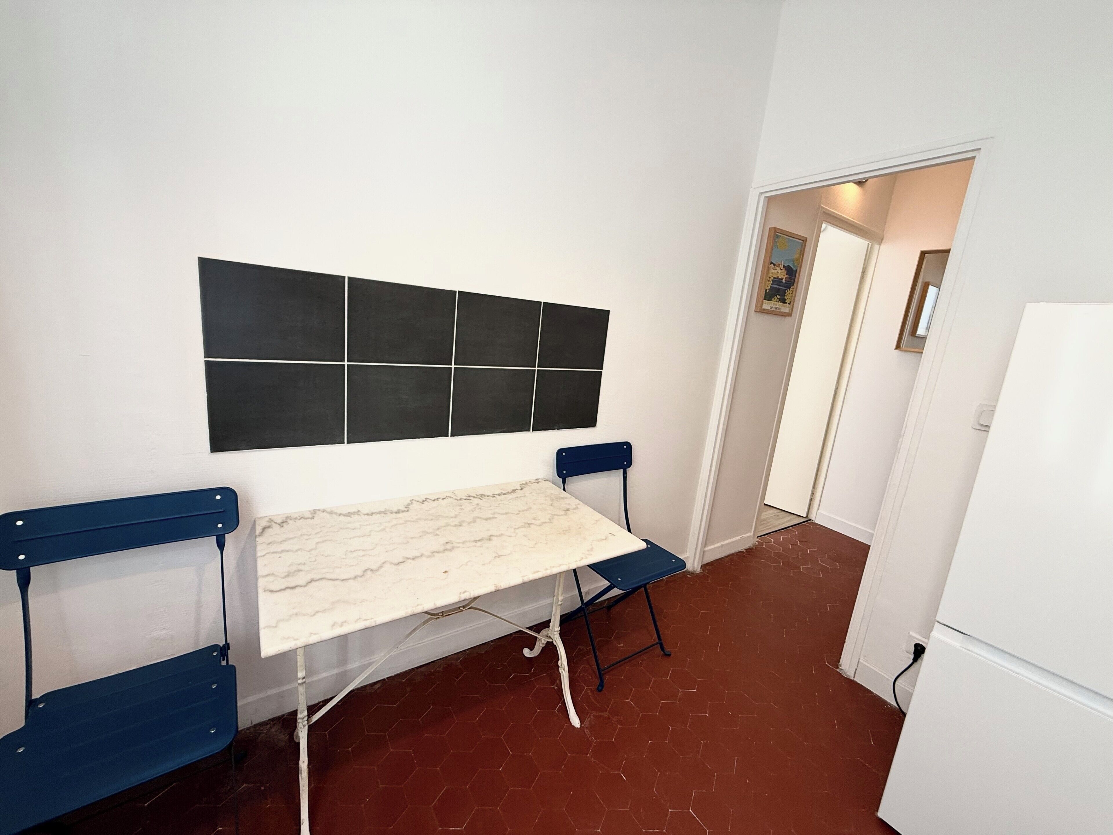 Une pièce avec un mur recouvert de carreaux blancs, une tête de lit en carreaux noirs, une table blanche avec un plateau imitant le marbre, et deux chaises bleues.