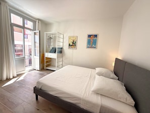 Una camera da letto con un letto, una scrivania e una sedia, un balcone vista edifici e due opere d'arte appese alle pareti.