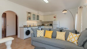 A compact living space with a grey sofa, a washing machine, a dining table, and a kitchen area. - Casa Oceanus - Magnificent Apartment with Pool (Cabanas de Tavira)