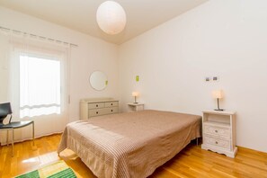 Een slaapkamer met een bed, een stoel, een dressoir, een nachtkastje, een lamp, een spiegel en een raam met gordijnen.