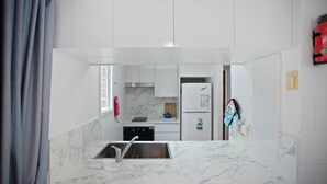 Una cocina compacta con armarios blancos, encimera de mármol, lavavajillas integrado y un ventanal pequeño.