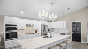 Une cuisine moderne avec des meubles blancs, un grand îlot central doté de tabourets, des appareils en acier inoxydable et un lustre.