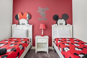 Une chambre avec deux lits, chacun recouvert d’une couette aux motifs de Mickey Mouse. Un mur rouge est décoré d’une silhouette de Mickey Mouse, et une petite table de chevet blanche avec une lampe se trouve entre les deux lits.