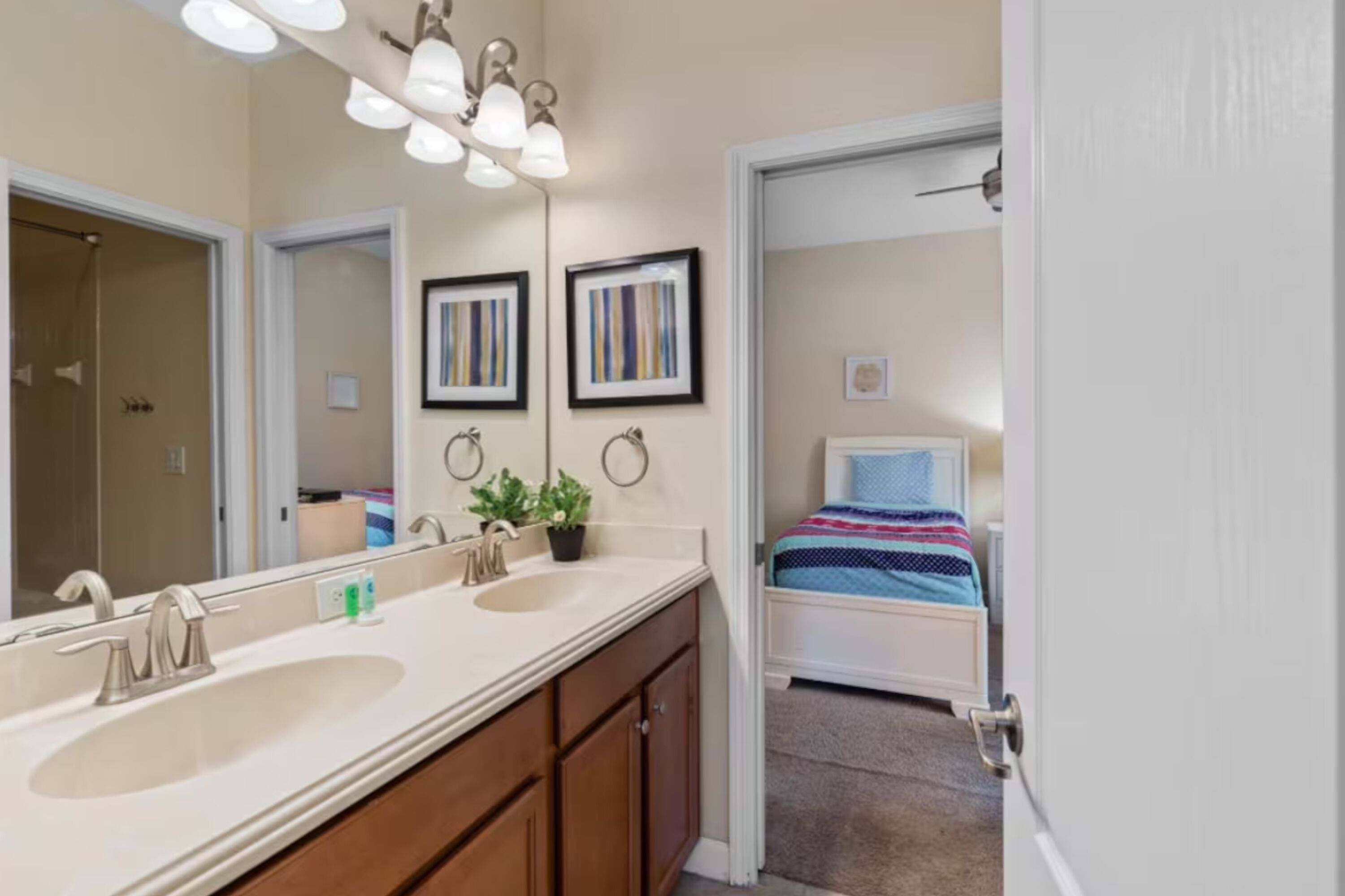A bathroom with a double sink, a mirror, and a shower. There is a bed with a blue and white striped bedspread in the adjacent room.