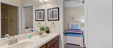 A bathroom with a double sink, a mirror, and a shower. There is a bed with a blue and white striped bedspread in the adjacent room.