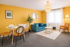 A modern living room with a blue sofa, a dining table with chairs, a small round table with a lamp, and a large window with curtains. - Manchester Apartment Free Parking (Salford)