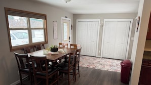 A dining area with a wooden table and chairs, a potted plant, and a rug. There are two white doors in the background.