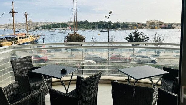 A balcony with wicker chairs and a table overlooking a marina with boats and a cityscape in the background. - Valletta Harbour View - Grand (Sliema)