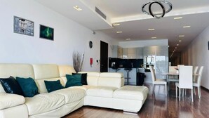 A modern living room with a beige sectional sofa, a dining area with white chairs, and a kitchen in the background. - Valletta Harbour View - Grand (Sliema)