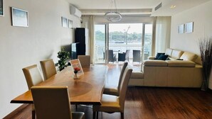 A modern living room with a wooden dining table, beige sofas, and a view of a body of water through large windows. - Valletta Harbour View - Grand (Sliema)