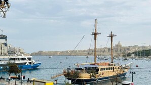 A harbor with a large sailing ship docked, a blue and white boat, and a cityscape in the background. - Valletta Harbour View - Grand (Sliema)