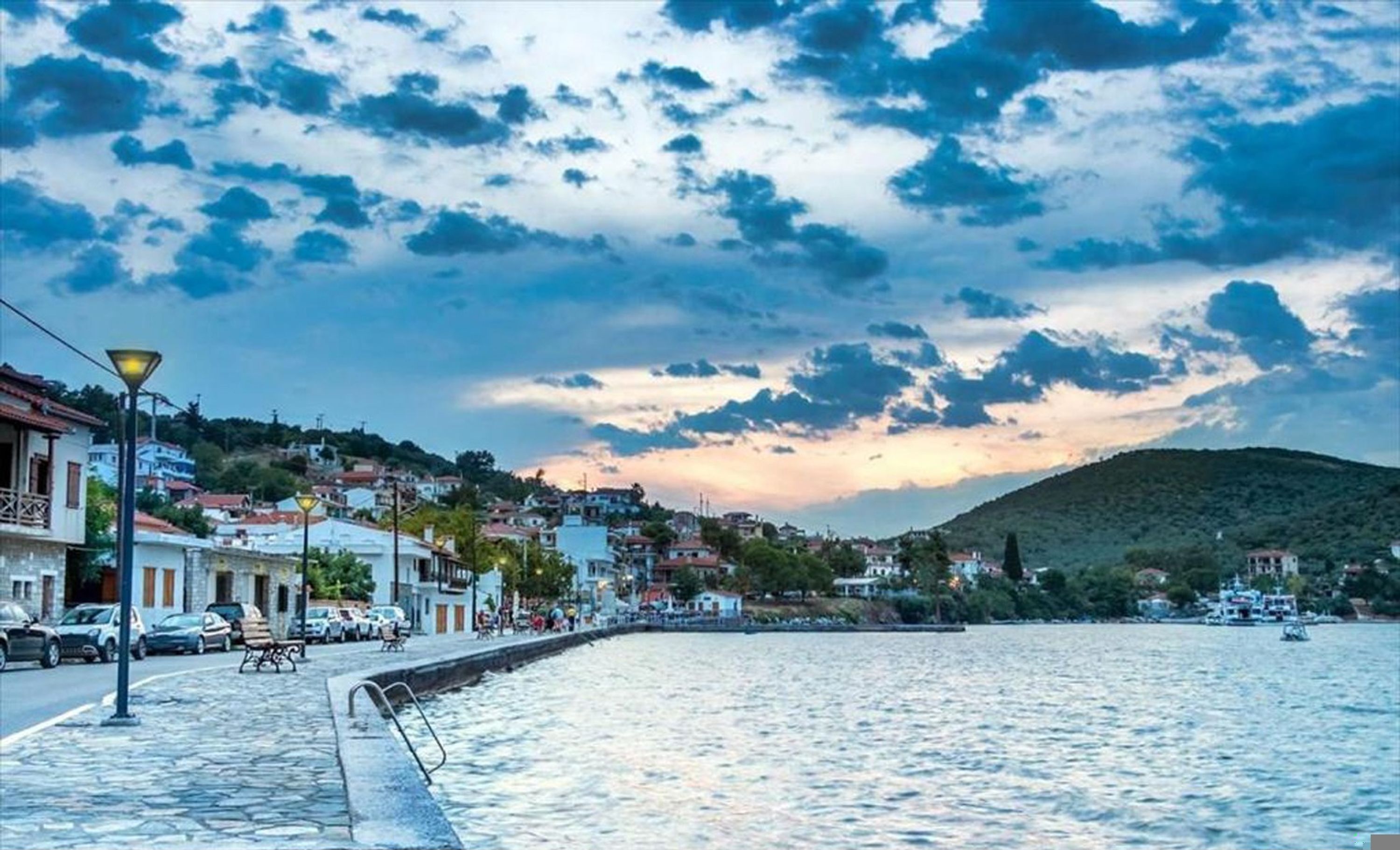 A coastal town with a stone-paved waterfront, buildings with balconies, and a hilly backdrop.
