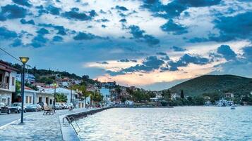 A coastal town with a stone-paved waterfront, buildings with balconies, and a hilly backdrop.