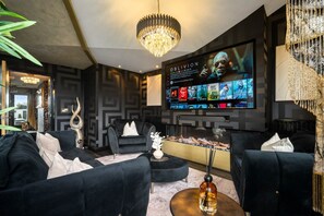 A modern living room with a large flat-screen TV displaying a movie, black sofas, a chandelier, and a decorative antler. - Opulent Penthouse - City Centre - 2Bedrooms - Balconies - 2Secure Parking Spaces (West Midlands)