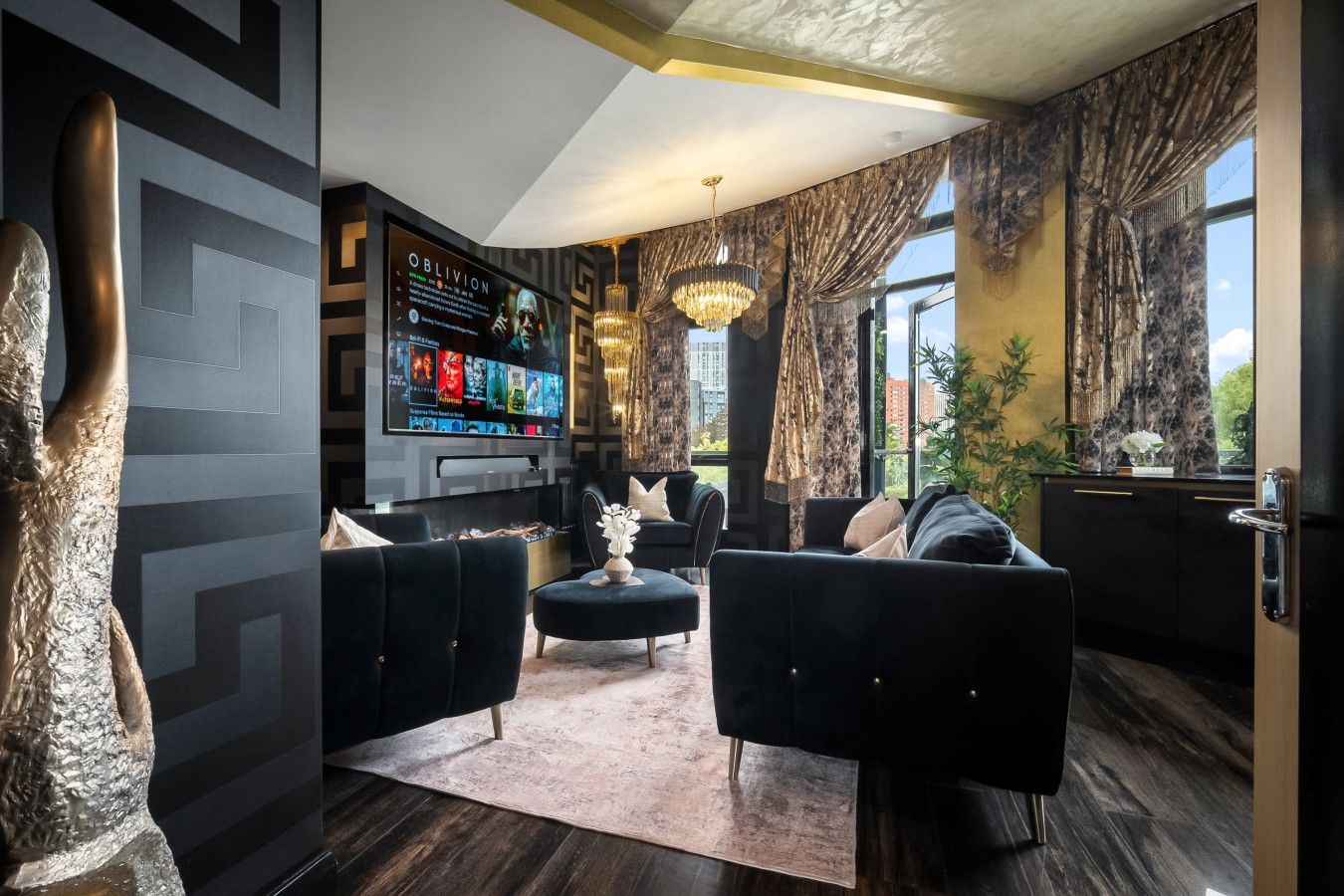 A modern living room with a flat-screen TV, dark sofas, a chandelier, and large windows.