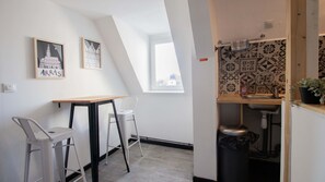 Eine kleine Küchenzeile mit Fenster, Spüle und Mülleimer. Zwei Stühle und ein Tisch mit einem Bilderrahmen an der Wand.