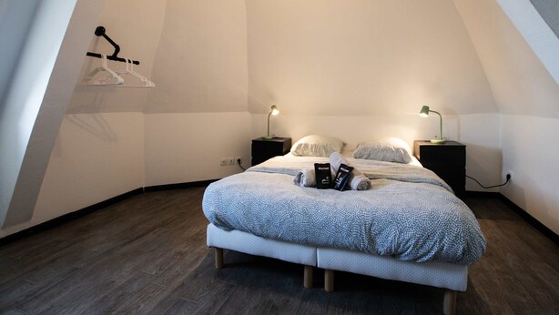 A bedroom with a sloped ceiling, a bed with a patterned duvet, two bedside tables with lamps, and a hanging clothes rack.