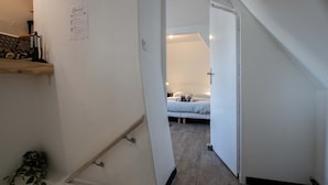 A narrow hallway leading to a bedroom with a bed, a bedside table, and a nightstand. There is a small shelf with decorative items on the left wall.