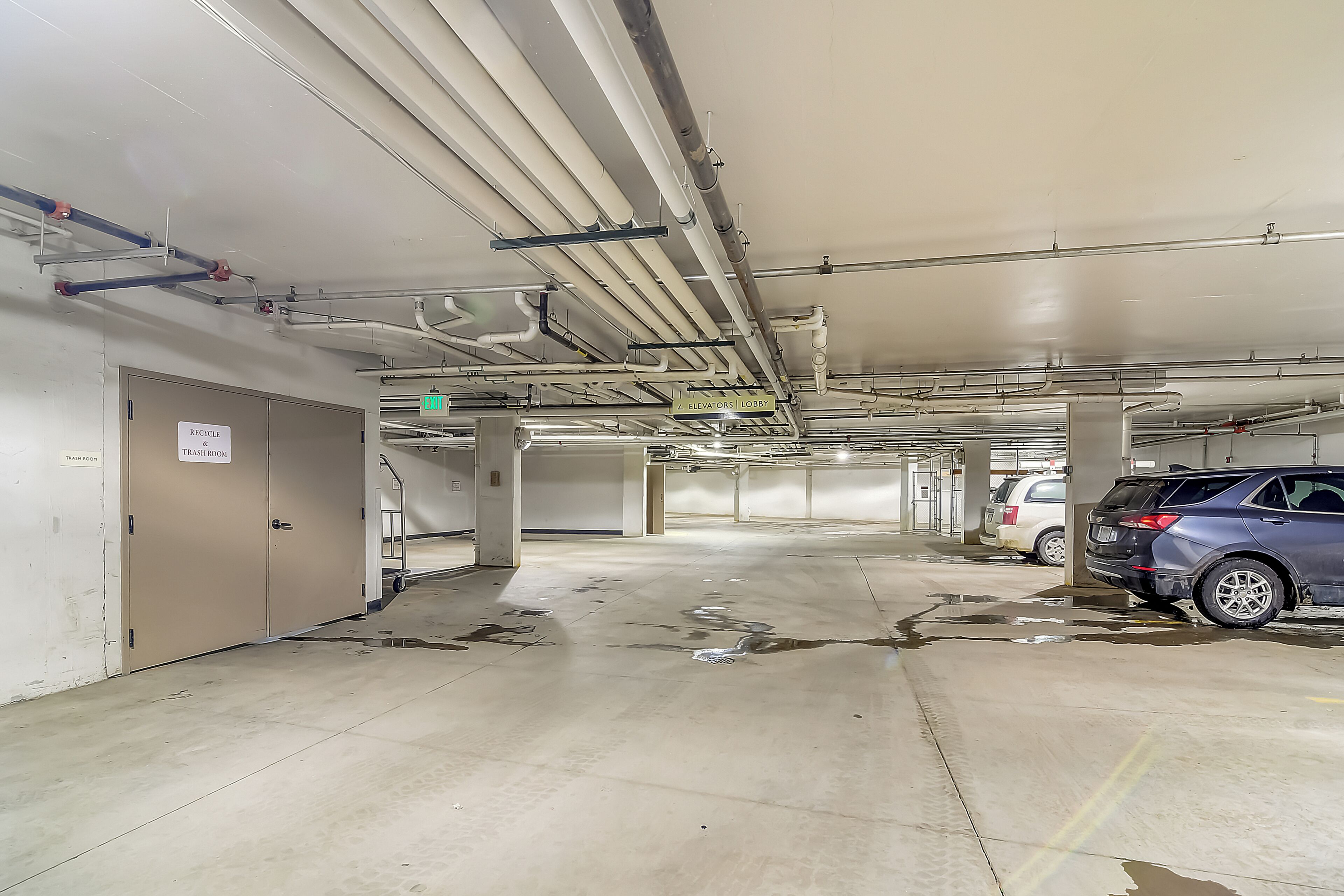 A parking garage with multiple levels, visible pipes and ducts on the ceiling, and a sign indicating an elevator lobby.