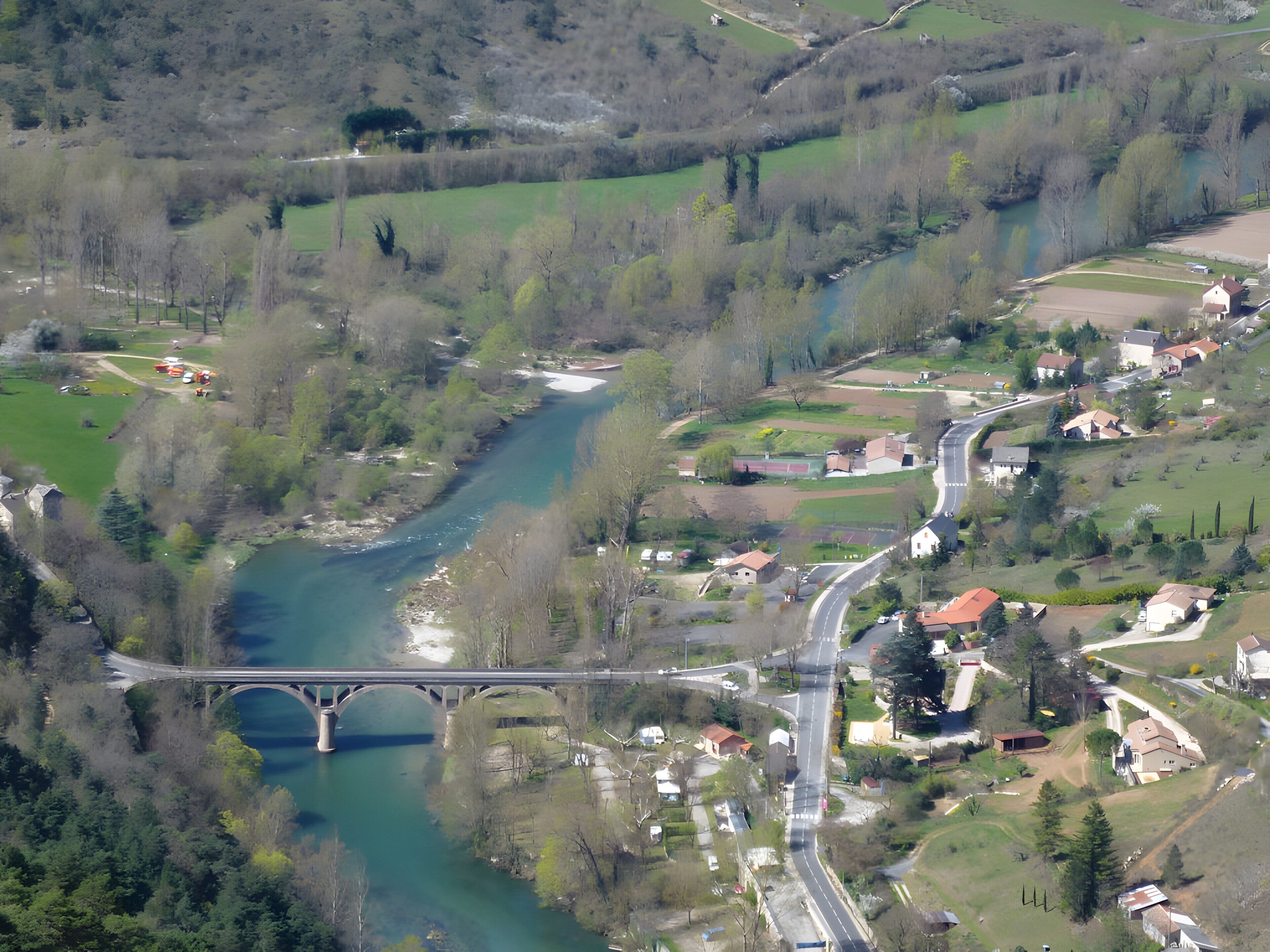 Vista aérea de uma área rural com um rio, uma ponte e algumas casas espalhadas.