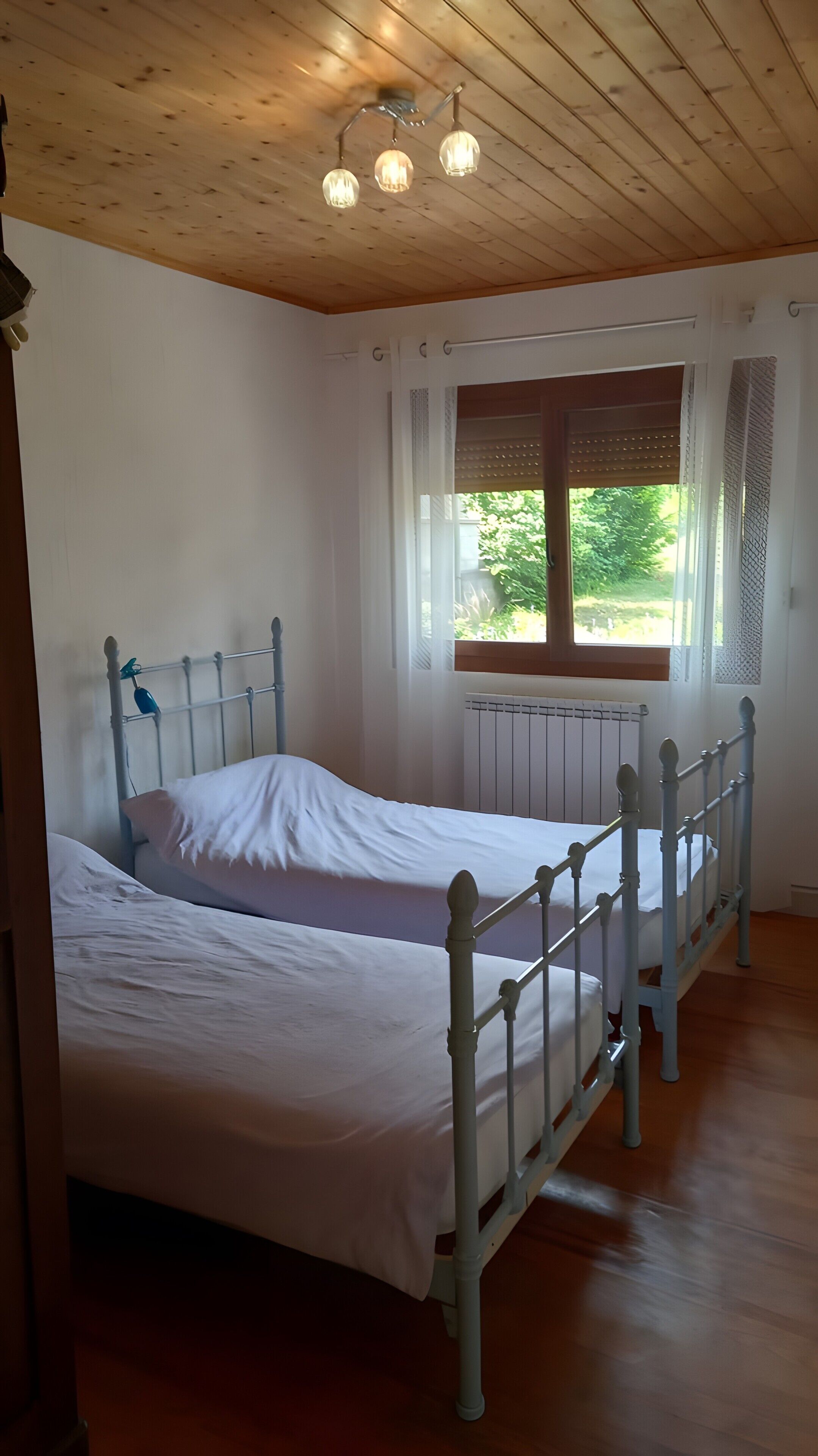 A room with a wooden ceiling, a bed with white bedding, a window with a view of greenery, and a radiator.