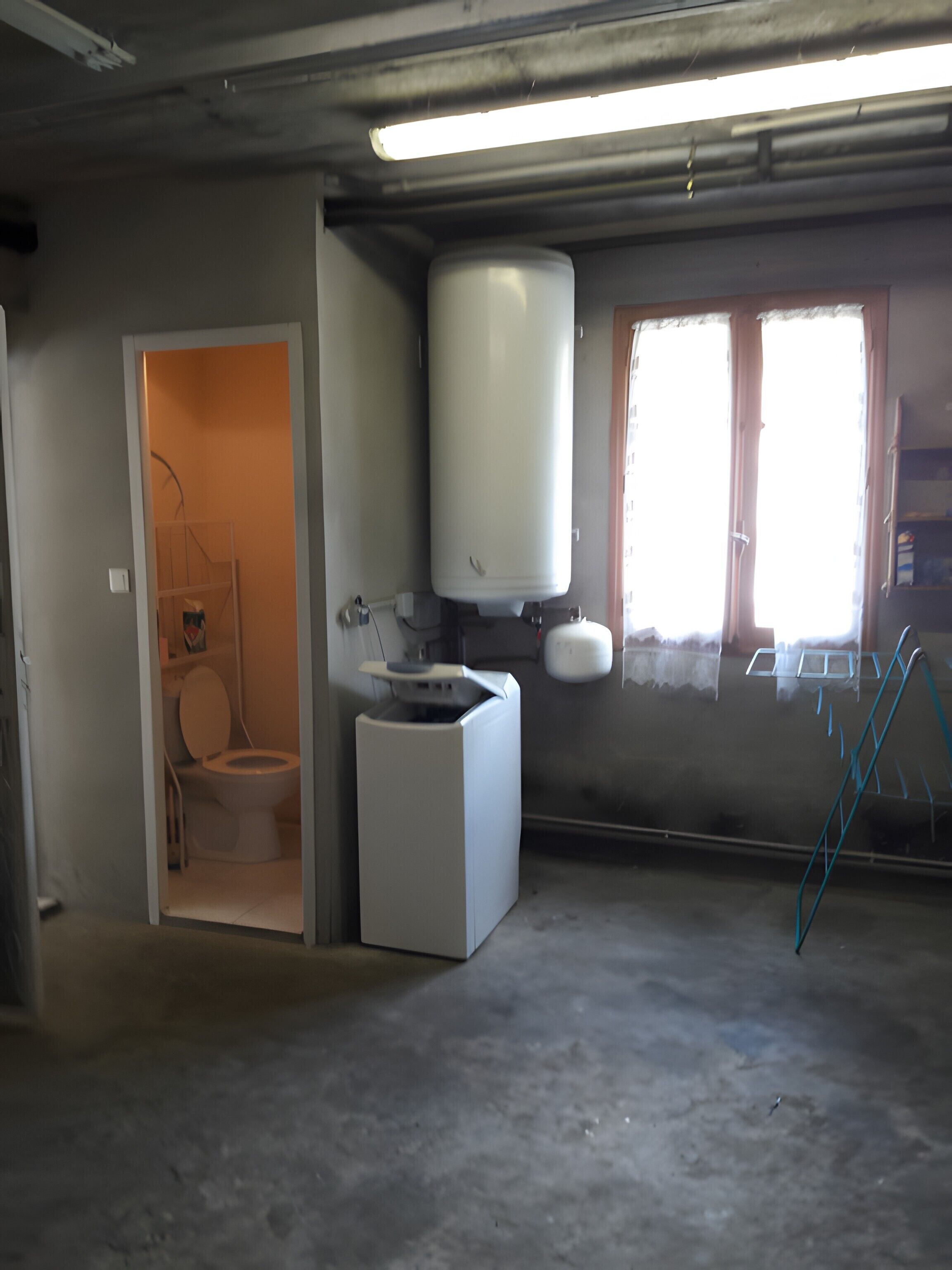 Une pièce équipée de toilettes, d’un lave-linge, d’un chauffe-eau et d’une fenêtre avec des stores.