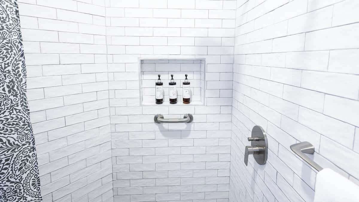 Un baño con paredes de azulejos blancos, zona de ducha y un estante con tres botellas.