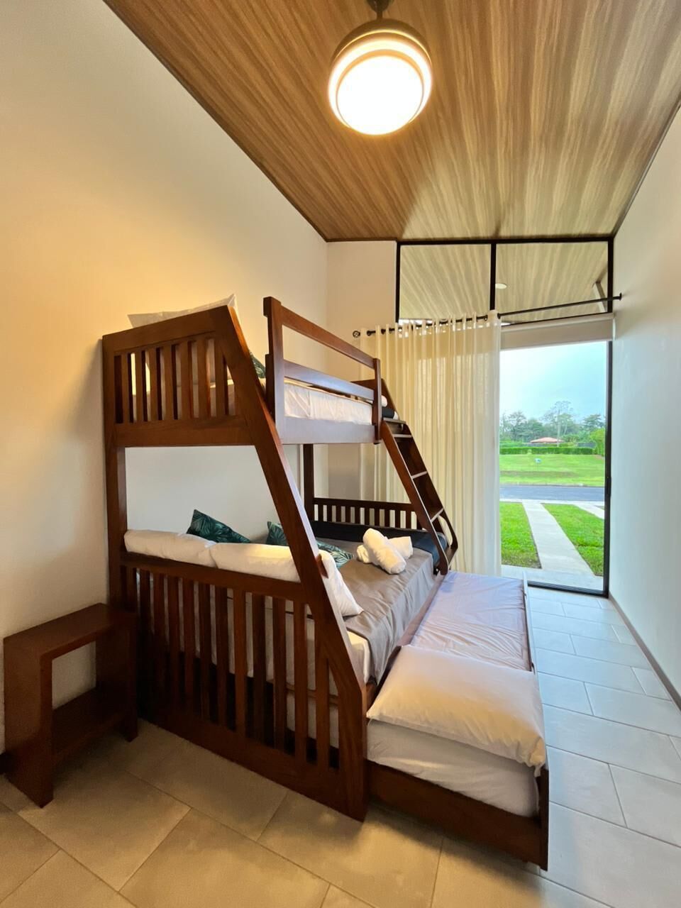 A bunk bed with a wooden frame, a bedside table, and a view of a green landscape through a window.