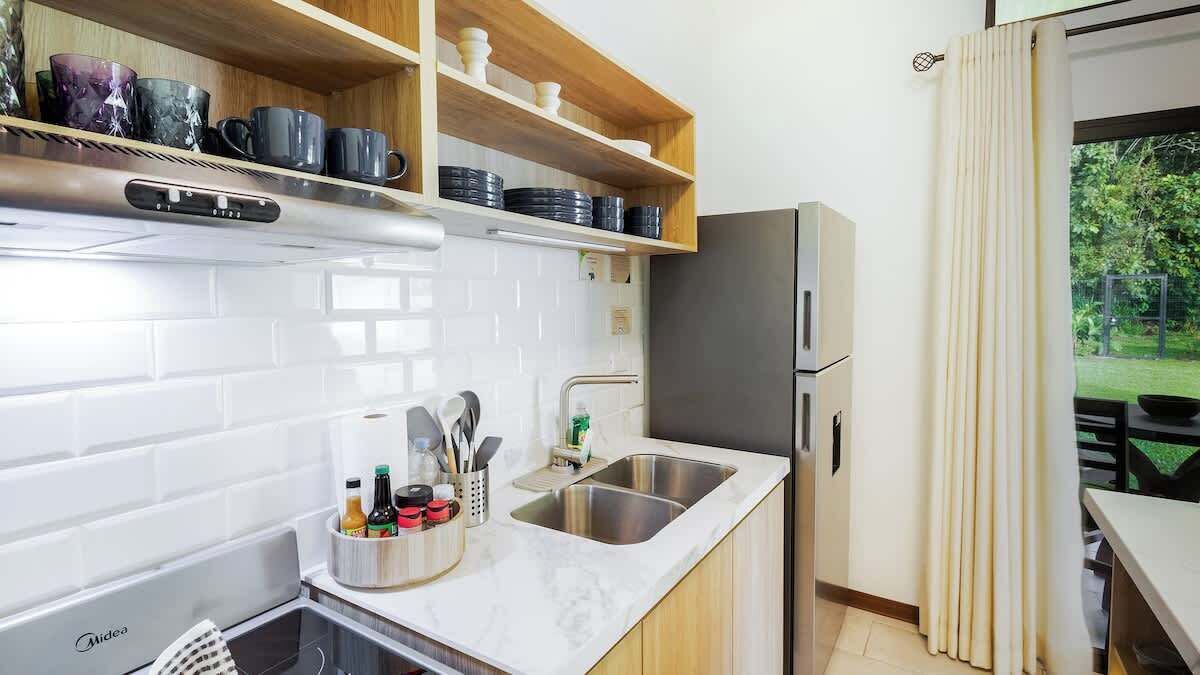 A kitchen with white tiled backsplash, stainless steel sink, wooden shelves with dishes, and a refrigerator.