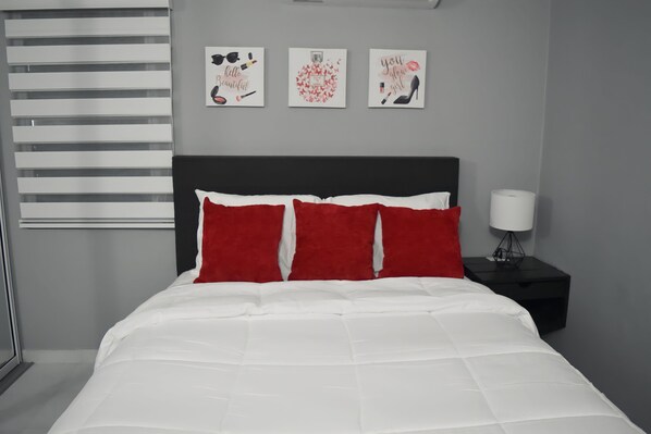 A bedroom with a bed, red pillows, a black nightstand with a lamp, and three wall art pieces.