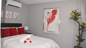 Een net opgemaakt bed met rode kussens, een ingelijst abstract schilderij en een pot met een plant.