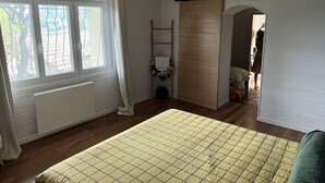 Quarto com cama, ventilador de teto, janela com cortinas, radiador e porta que dá para outro cômodo.
