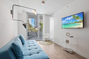 A modern living room with a blue sofa, a mounted TV displaying a beach scene, a clear glass side table, and a balcony with a view of buildings and palm trees. - Beach Escape 1BR • Walk to beach • Pool & Gym (Miami Beach)