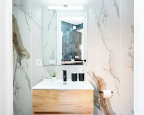 Une salle de bain avec un mur en marbre, un lavabo blanc, un meuble en bois, un miroir et un pare-chocs en carreaux bleus.