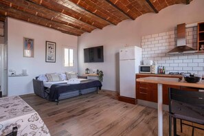 Ein kleines Zimmer mit Bett, einer kompakten Küchenzeile und einem an der Wand befestigten Fernseher.