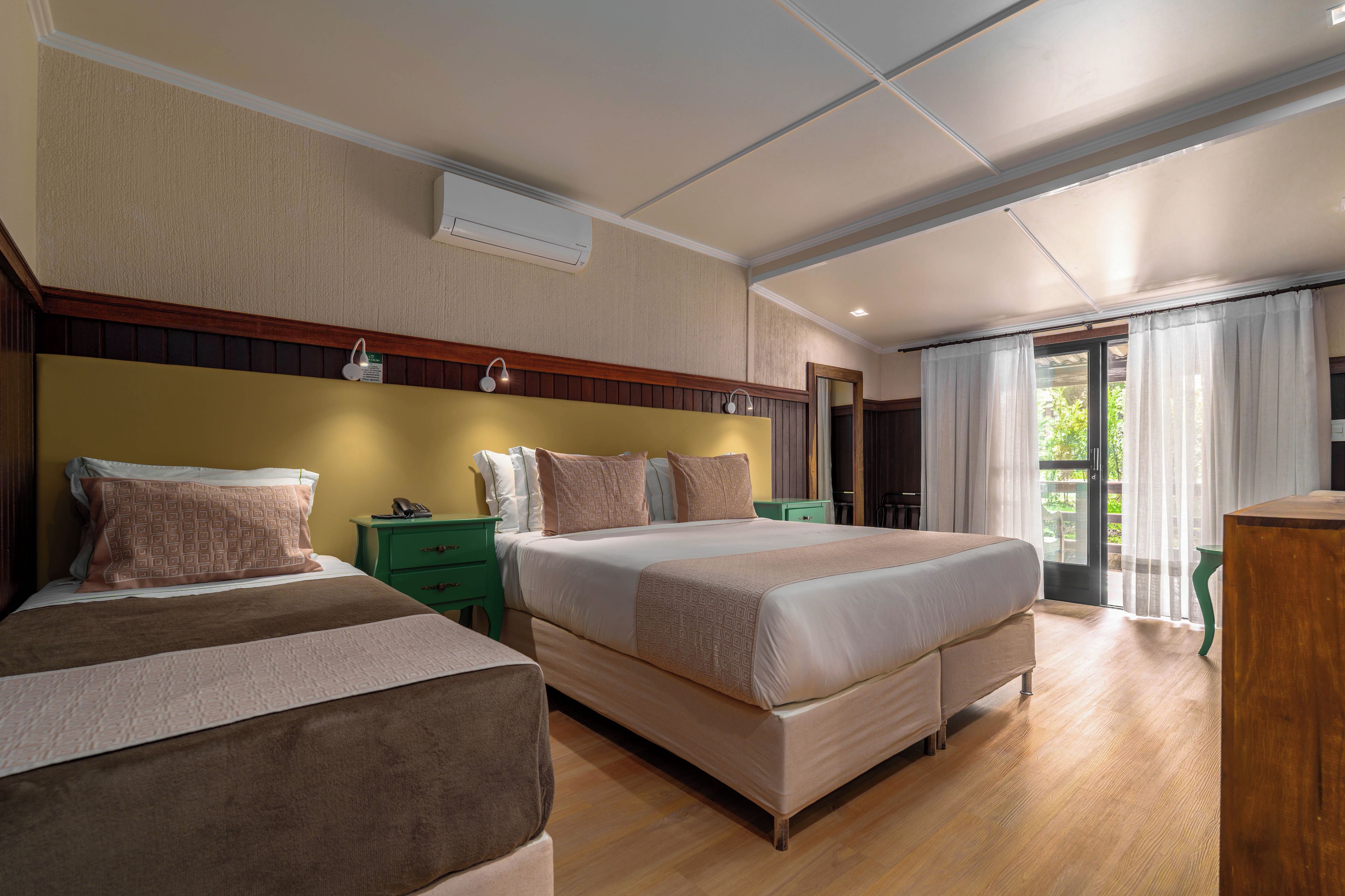 Een hotelkamer met twee bedden, een groen nachtkastje, een houten hoofdbord, een groot raam met gordijnen en een plafondlamp.