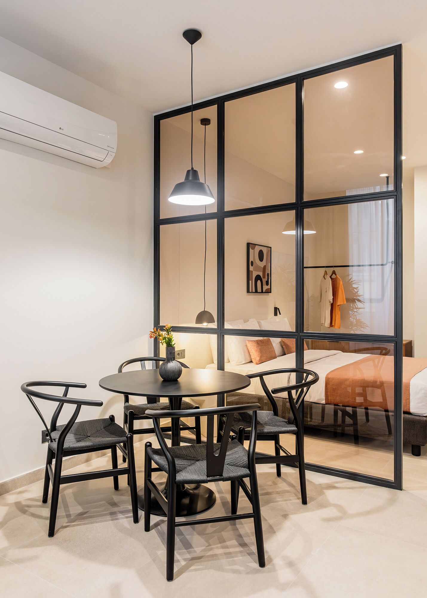 A modern room with a round table, four chairs, a hanging pendant light, and a large window with a view of a bed and a plant.